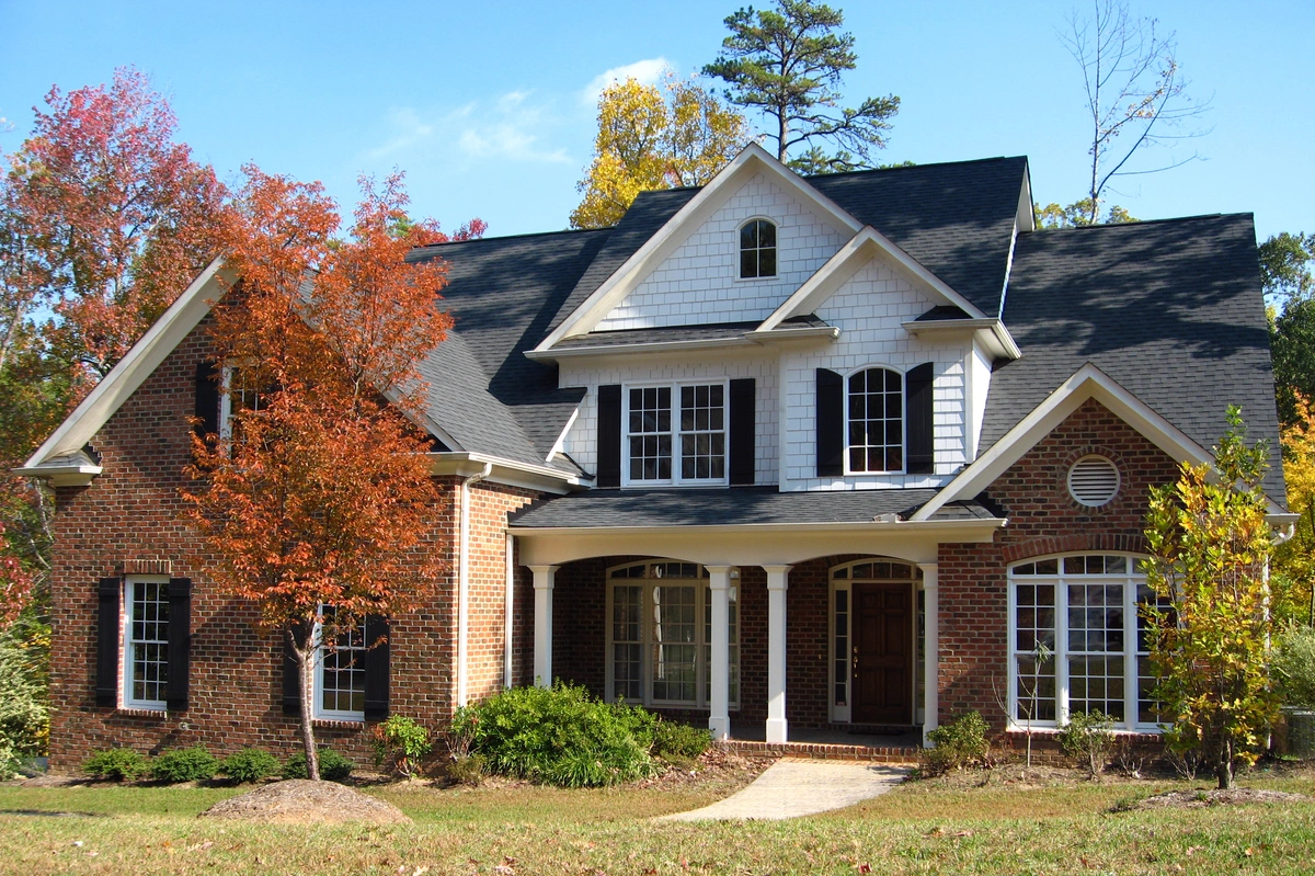 A two-story brick house with white trim and a dark shingled roof, expertly installed by a local roofing company, is surrounded by trees with autumn foliage. The home features large windows, a gabled front entrance, and a well-maintained lawn.