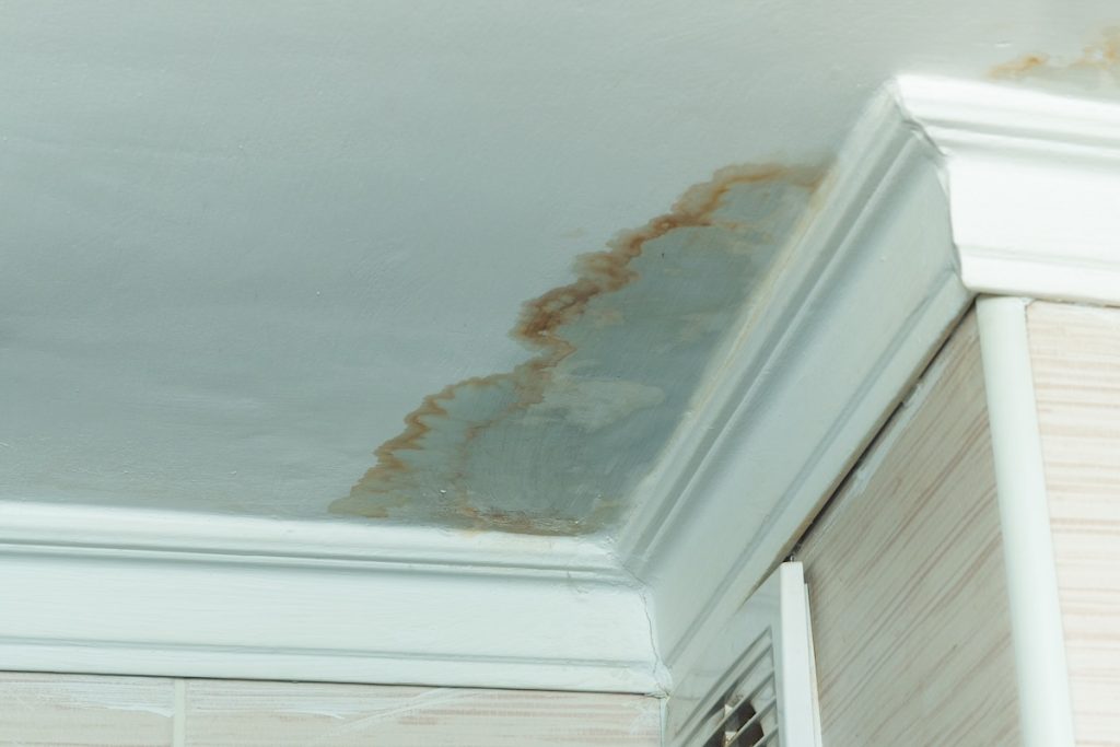 A ceiling corner with visible water damage, likely from a leak, shows brown stains and discoloration near the white crown molding above tiled walls—signs that may require a storm damage roof inspection.