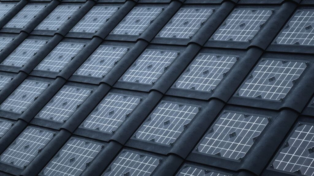 Close-up view of a slanted roof with integrated solar panels, each panel framed by dark roof tiles in a grid-like pattern. The panels feature visible grid lines and are set among different types of roof shingles.