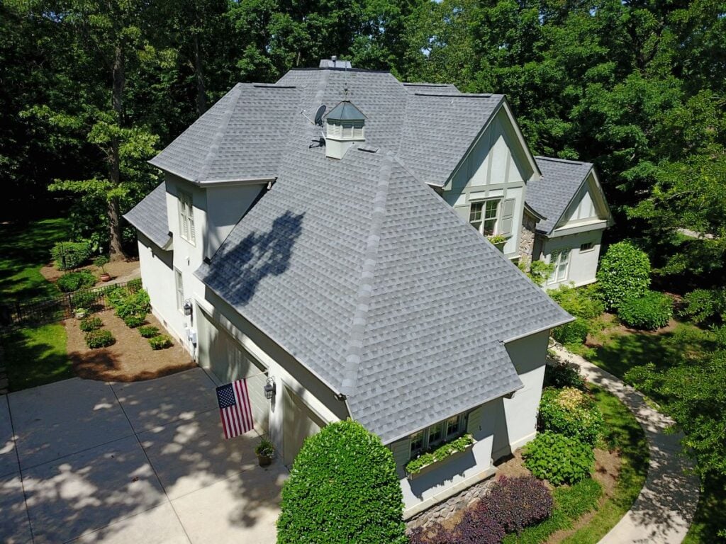 Aerial view of a large modern house with a grey shingle roof, white exterior walls, multiple gables, and a driveway. The well-landscaped home hints at quality features throughout—imagine investing in curb appeal or considering future roof replacement cost.