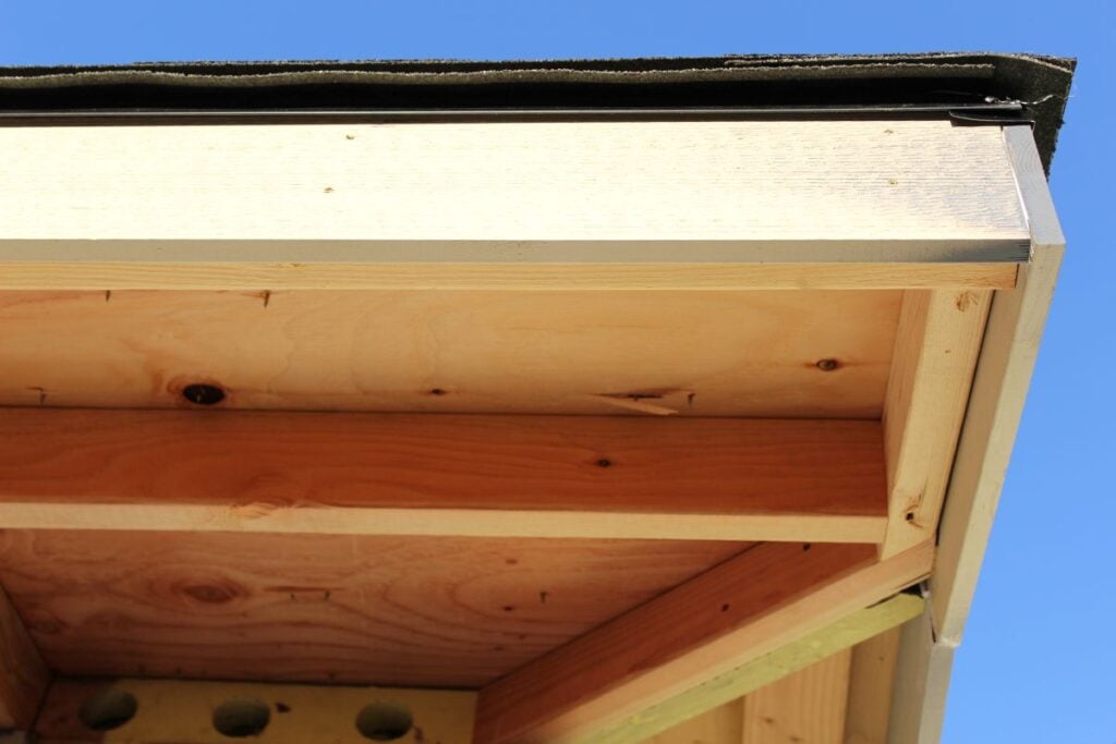 Close-up view of the eaves of a house under construction, showing exposed beams, light-colored wood, and a clear blue sky in the background.