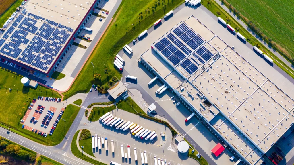 Aerial view showcasing two distinct commercial roof types: one large, flat roof covered extensively with solar panels, and another large, flat roof with a gravel surface, both on industrial buildings with loading docks.