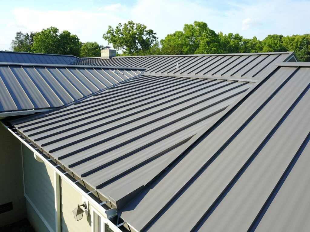A close-up view of a modern house’s gray standing seam metal roof, showcasing sleek panels and surrounded by green trees under a partly cloudy sky, hints at the quality and value reflected in standing seam metal roof cost.