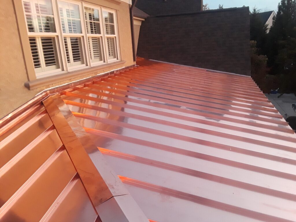 A shiny copper standing seam metal roof, known for its durability and premium standing seam metal roof cost, is attached to a tan house with white-framed windows and shutters, reflecting the evening light.