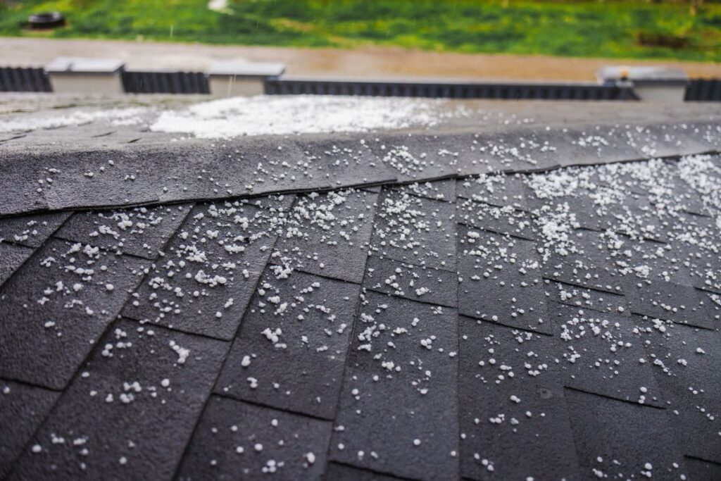 Small white hailstones are scattered across dark asphalt roof shingles, hinting at potential hail damage to the roof, with a blurred background of green grass and pavement visible beyond the edge of the roof.