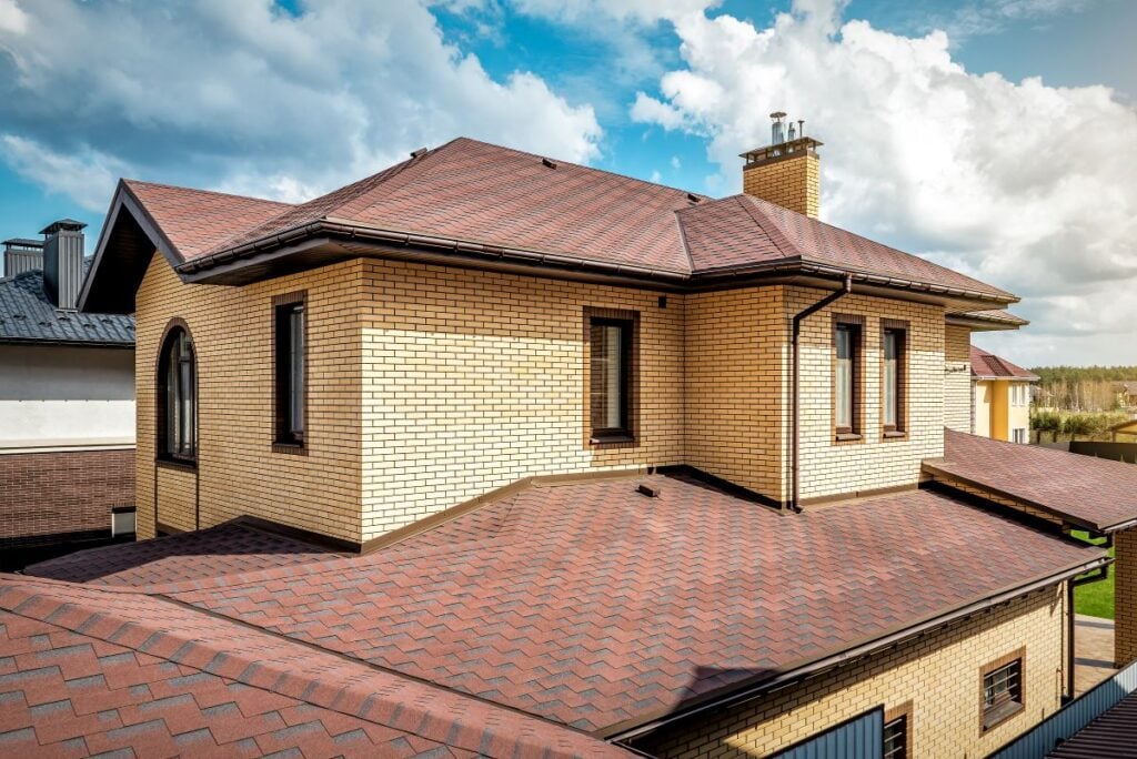 house and roof color combinations Two-story house with light yellow brick walls and a brown shingle roof under a partly cloudy sky. The harmonious house and roof color combinations are highlighted by the warm yellow bricks paired with rich brown shingles. The roof features multiple slopes and a chimney, with several windows visible on both floors.