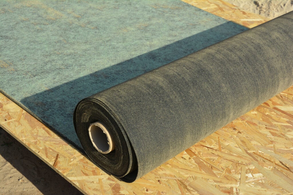 A roll of black roof felt underlayment partially unrolled on a sheet of oriented strand board.