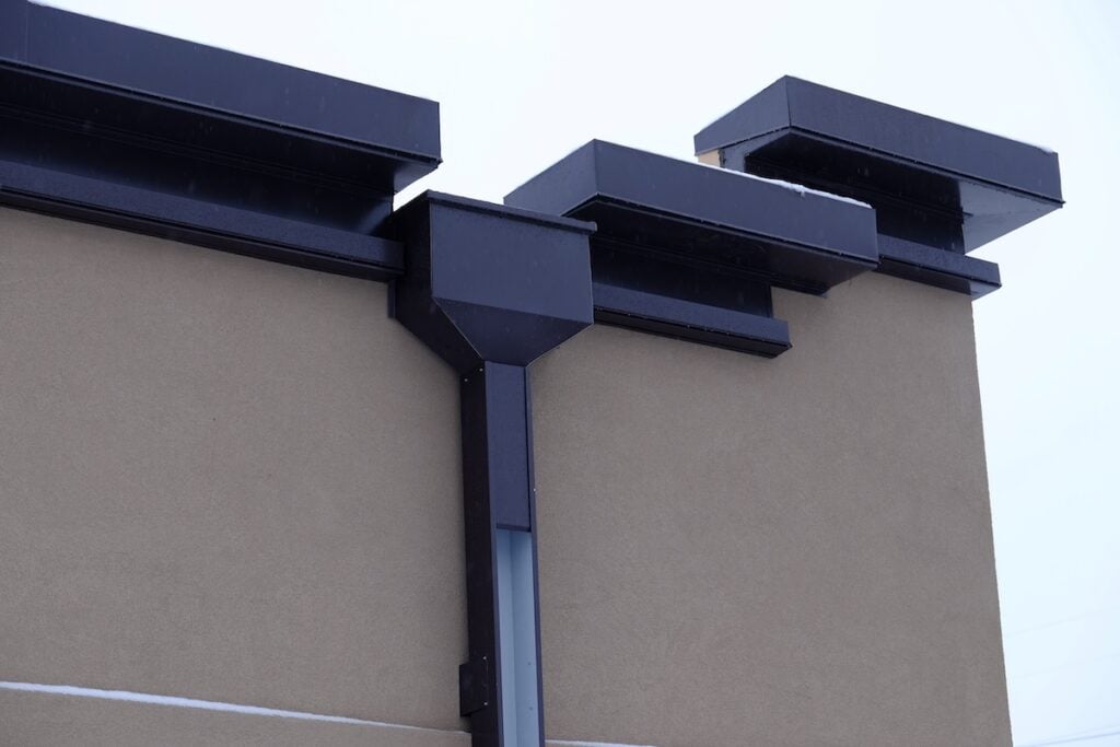 The image shows a modern building roof with three rectangular, black vents or chimneys extending from a beige wall. The vents are arranged in a row, with clean lines and a minimalist design against a cloudy sky.