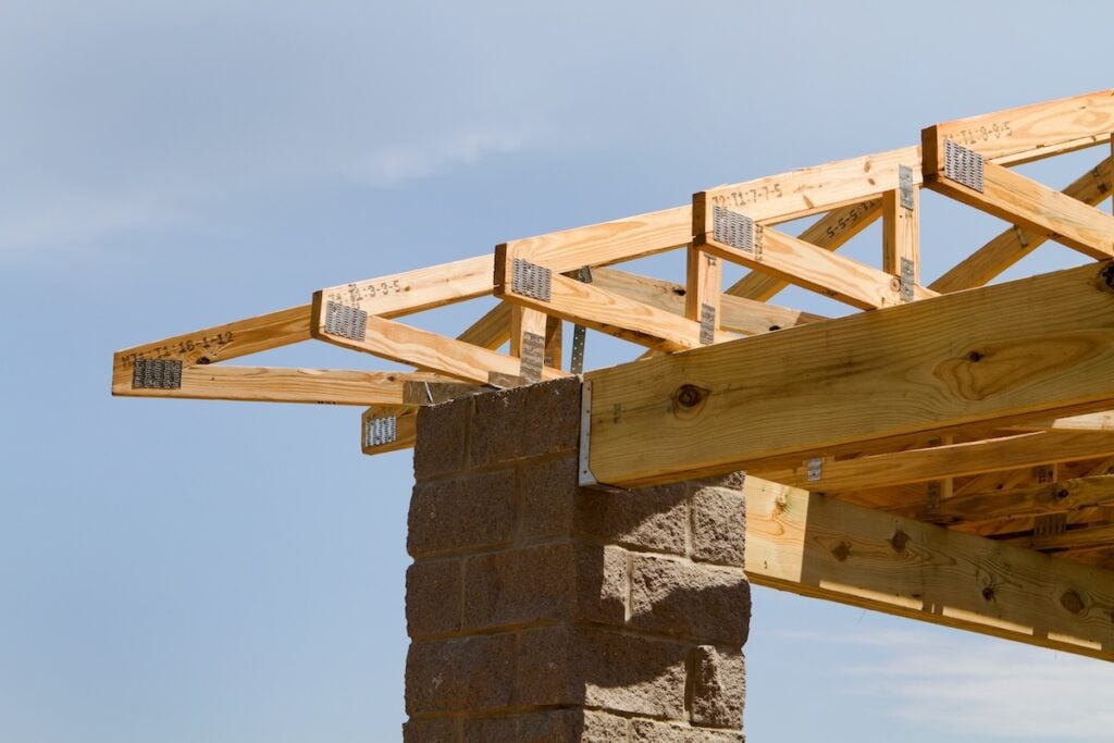 Close-up of wooden roof trusses being constructed on top of a brick pillar against a clear blue sky. The trusses are fastened with metal connectors and include measurements and markings on the wood.