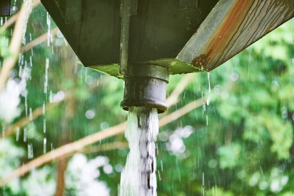 Close-up of water flowing from a pipe attached to a structure, set against a blurred background of green trees. The image shows the motion of the water as it spills out, suggesting rain or drainage.