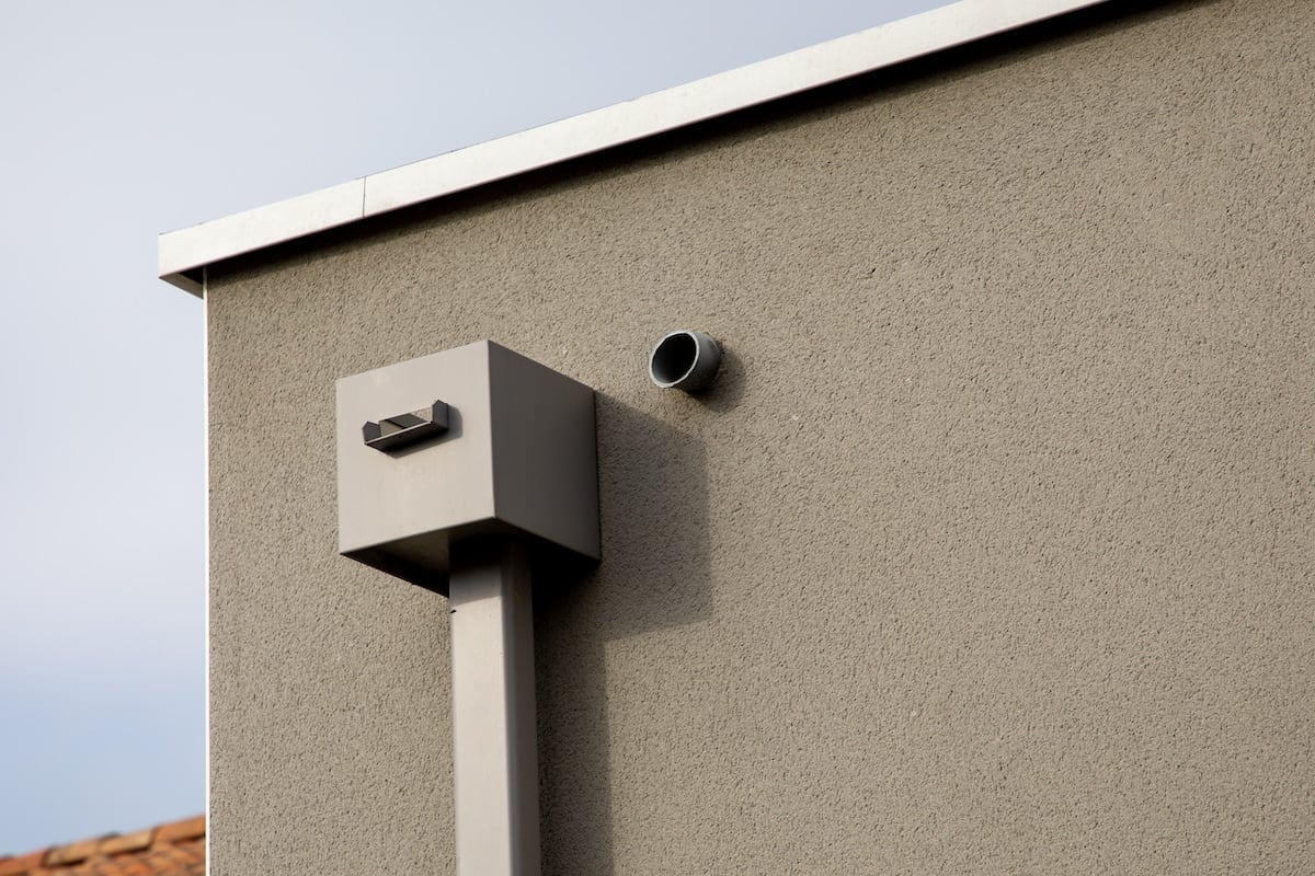 A minimalist exterior of a beige building wall featuring a small protruding metal vent pipe and a larger, boxy structure attached to a vertical pipe. The sky is overcast, and part of a tiled roof is visible in the corner.