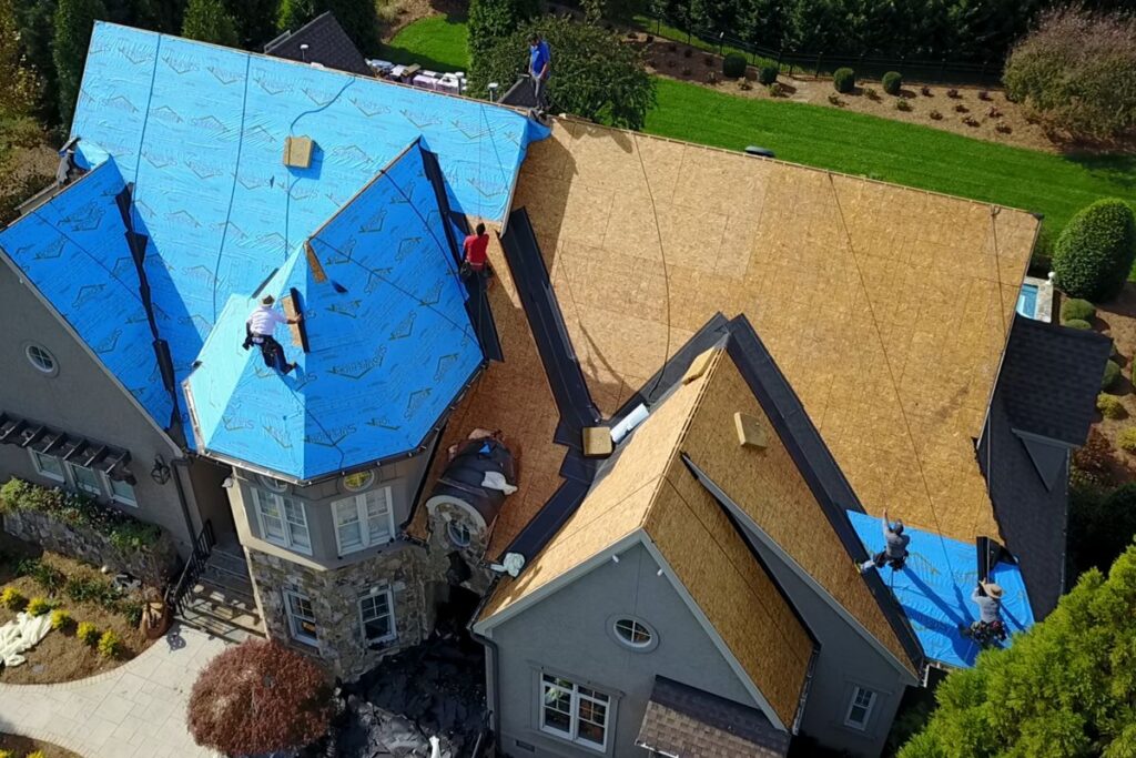 Aerial view of a house being roofed, with workers installing blue underlayment on the wooden roof sheathing. The house is surrounded by greenery, and workers are spread across different roof sections.