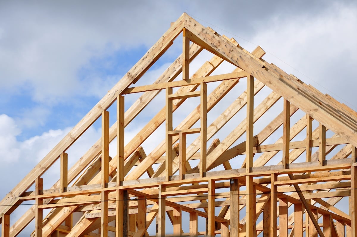 Wooden framing of a house under construction, showing the triangular roof structure against a cloudy sky background. The beams and joists form a grid-like pattern, illustrating the initial stages of building.