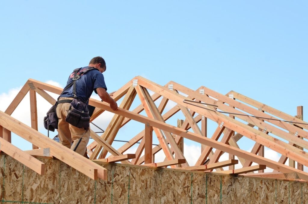 A construction worker in a blue shirt and tool belt stands on a wooden frame, building a roof on a house. The structure is made of unfinished timber beams. The sky is clear and blue in the background.