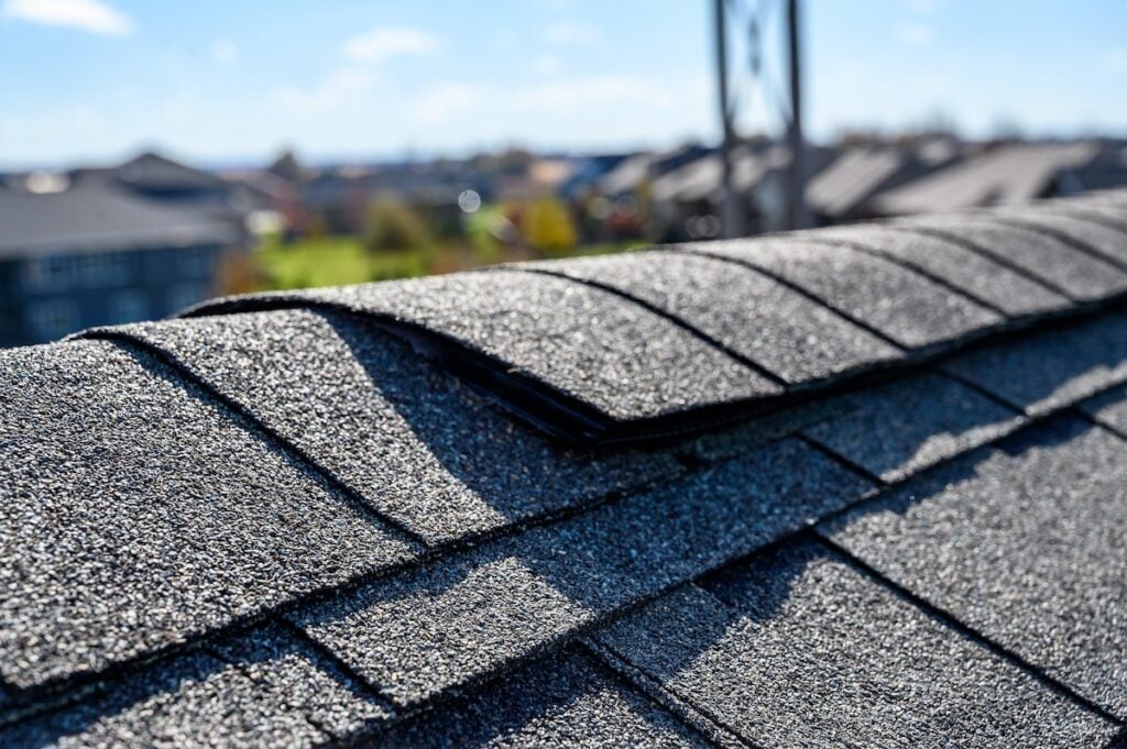 Close-up of a roof with asphalt shingles, focusing on a raised shingle along the ridge. The background shows blurred rooftops and a clear blue sky, suggesting a suburban setting.