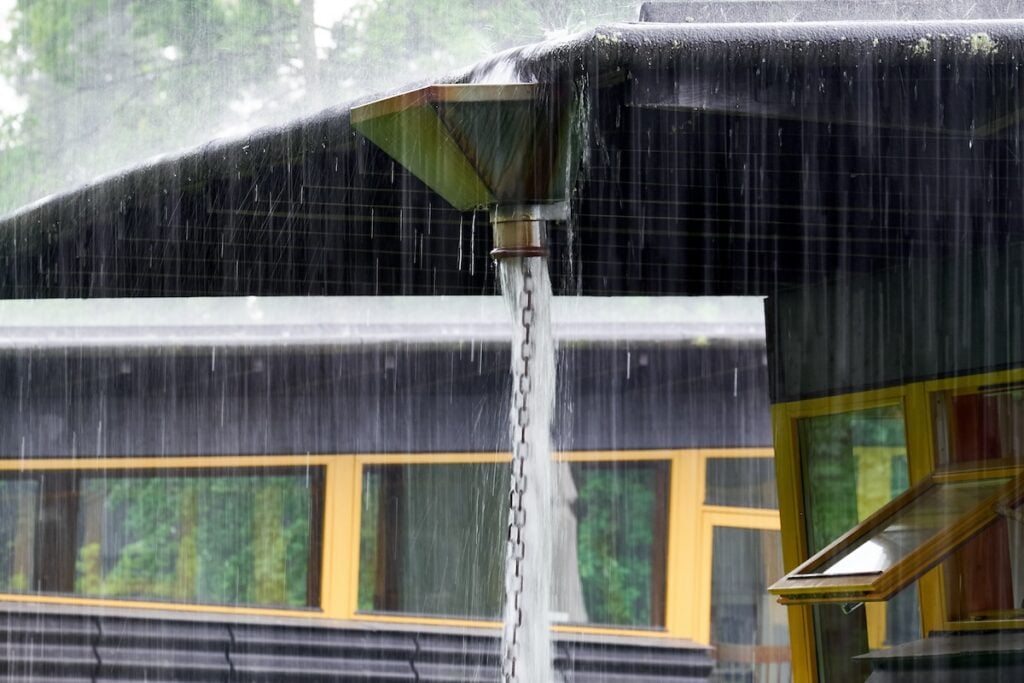 Rainwater pours from a slanted roof into a funnel, guided down by a metal chain. A modern building with yellow-framed windows is in the background, surrounded by greenery.