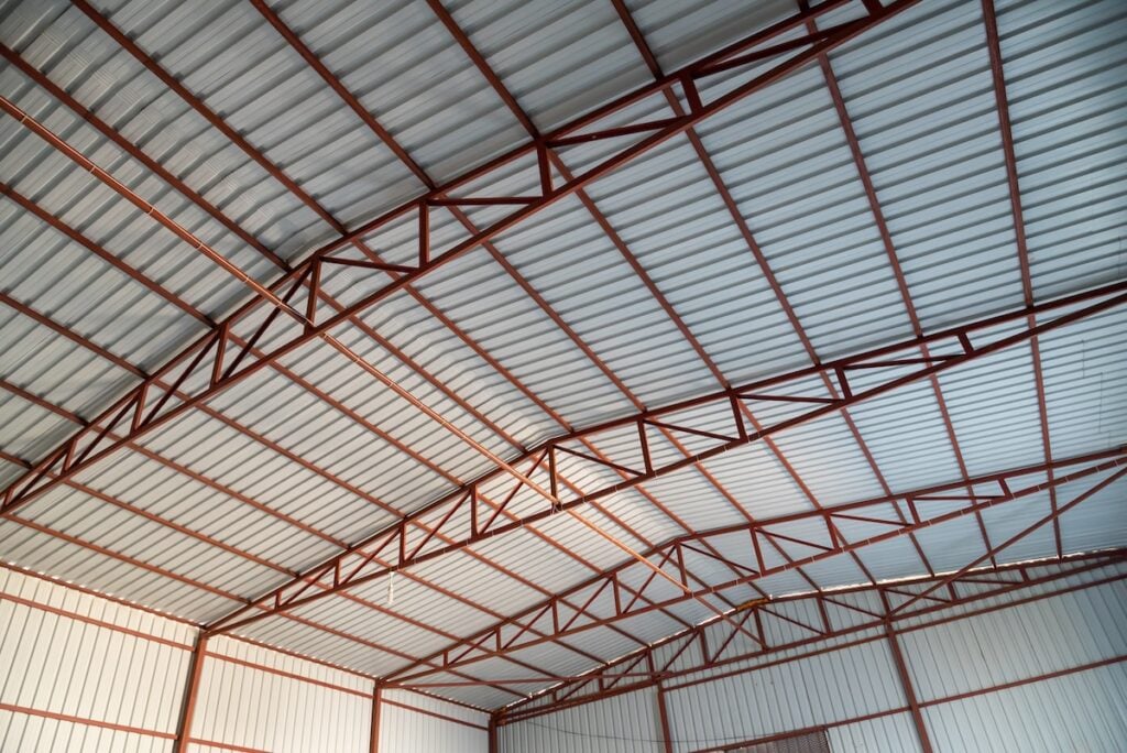Interior view of a large warehouse ceiling with corrugated metal sheets and red steel trusses. The roof structure creates a geometric pattern of intersecting lines and beams. The lighting highlights the roof's industrial design.