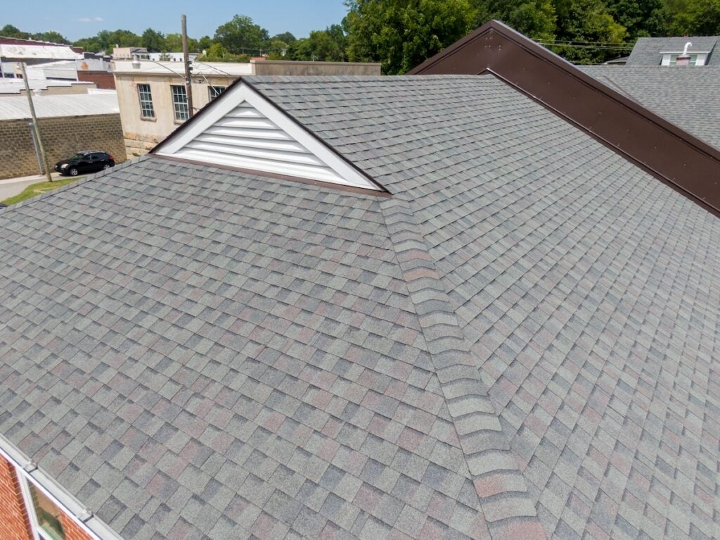 A rooftop with overlapping gray shingles, arranged in a diamond pattern, with a triangular attic vent on one section. Surrounding the roof are buildings and trees against a clear blue sky.