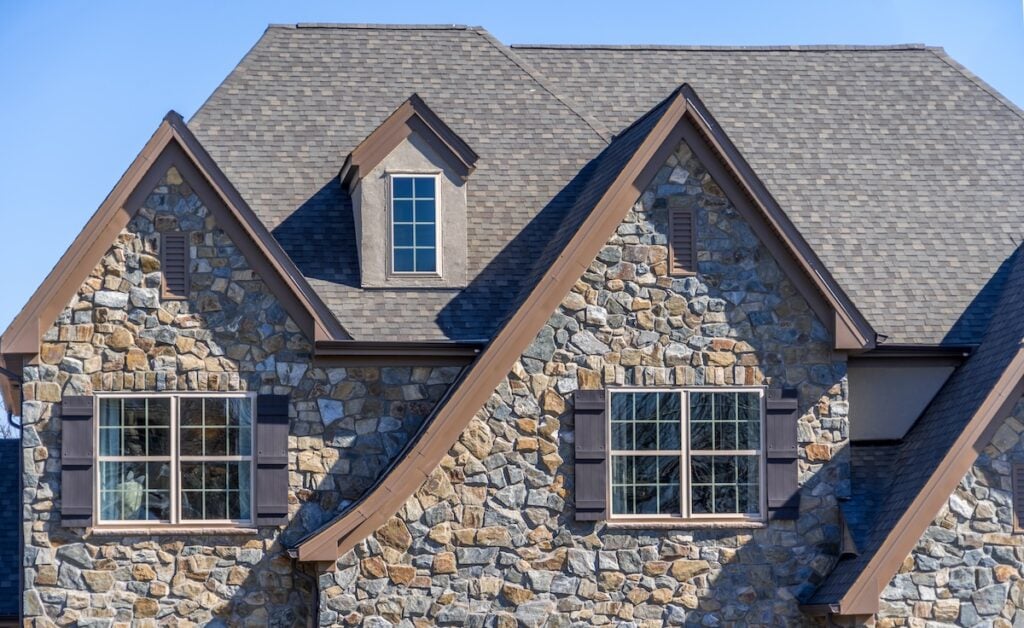 A close-up view of a stone house with a steeply pitched roof. The building features three windows, two on the lower level with shutters and a smaller one near the roof peak. The shingles and stones have earthy tones durham