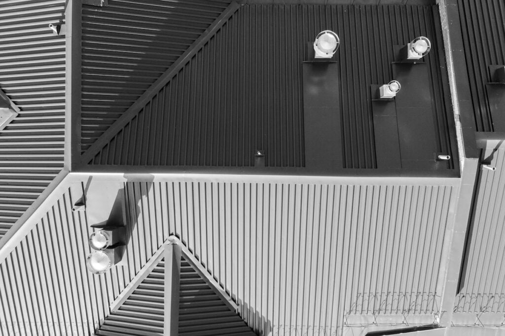 wake forest nc commercial Black and white image of a modern roof in Lake Forest, featuring triangular sections and corrugated metal sheets. Cylindrical lights are mounted on the slanted surfaces, creating geometric patterns. A section of chain-link fence is visible at the edge.