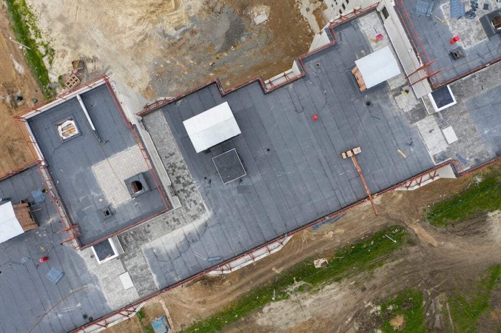 Aerial view of a flat-roofed building under construction in Lake Forest. The roof is mostly covered in black material, with some white structures and metal frames visible. Surrounding the site are patches of dirt, grass, and various construction materials scattered around. wake forest nc