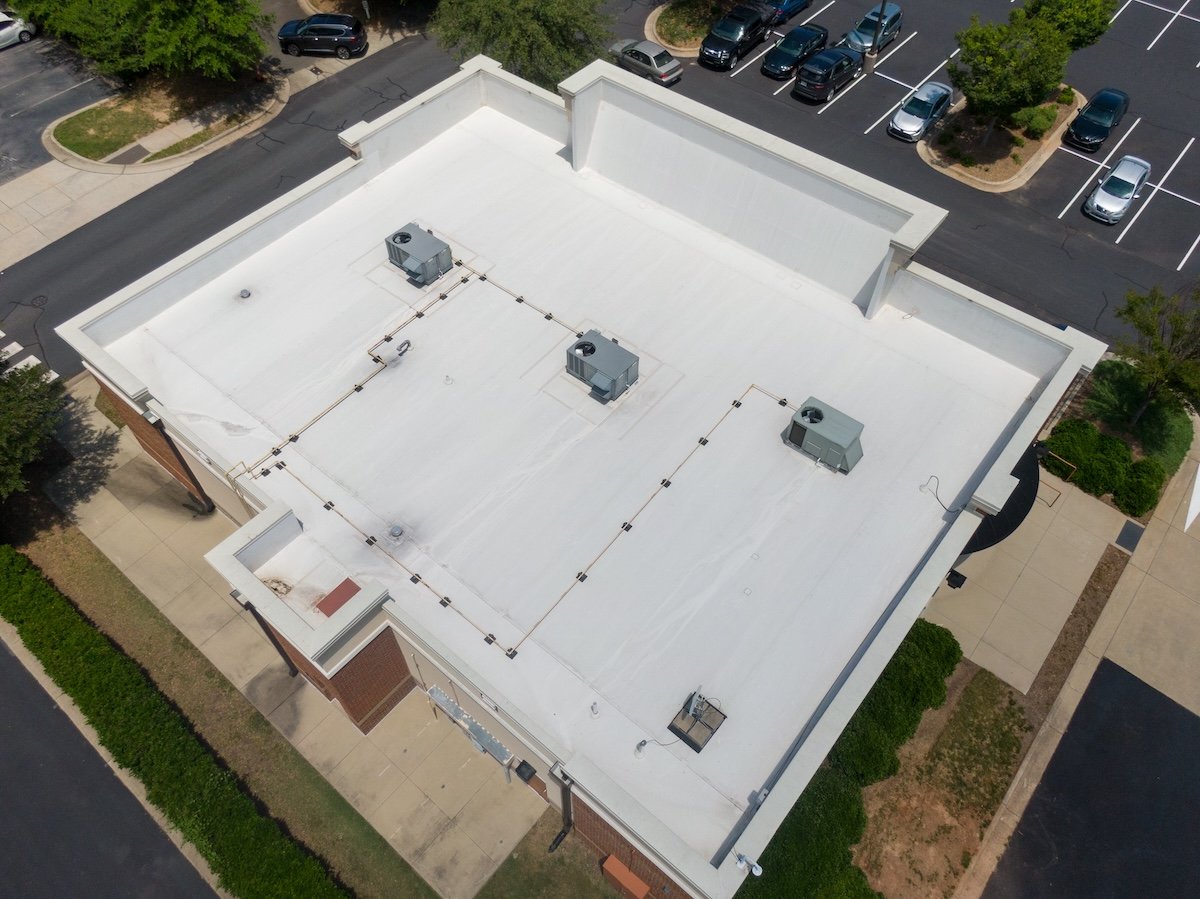apex nc commercial Aerial view of a flat rooftop with a white surface, featuring three air conditioning units and a few pipes. This pristine roofing in Lake Forest is surrounded by parking spaces filled with several cars and landscaped areas brimming with lush greenery.