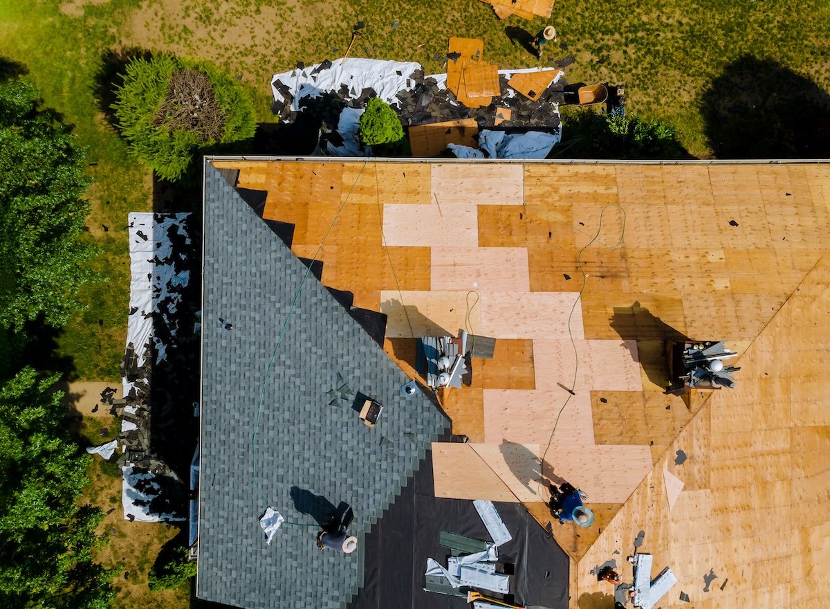 Aerial view of a house roof undergoing renovation in apex. Part of the roofing is covered with green shingles, while wooden panels and tarps expose another section. Construction materials and tools are scattered across the roof, surrounded by lush greenery. north carolina