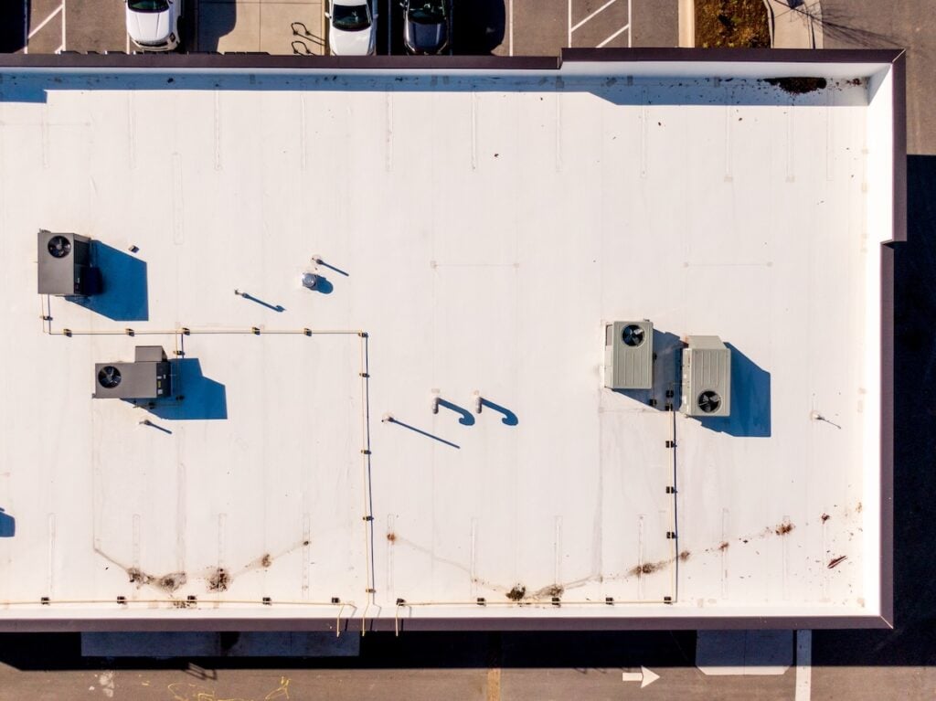 Aerial view of a flat, white morrsiville nc commercial building roof with HVAC units and various pipes and vents casting shadow outlines. Edges are visible, bordered by a parking lot with some parked cars.