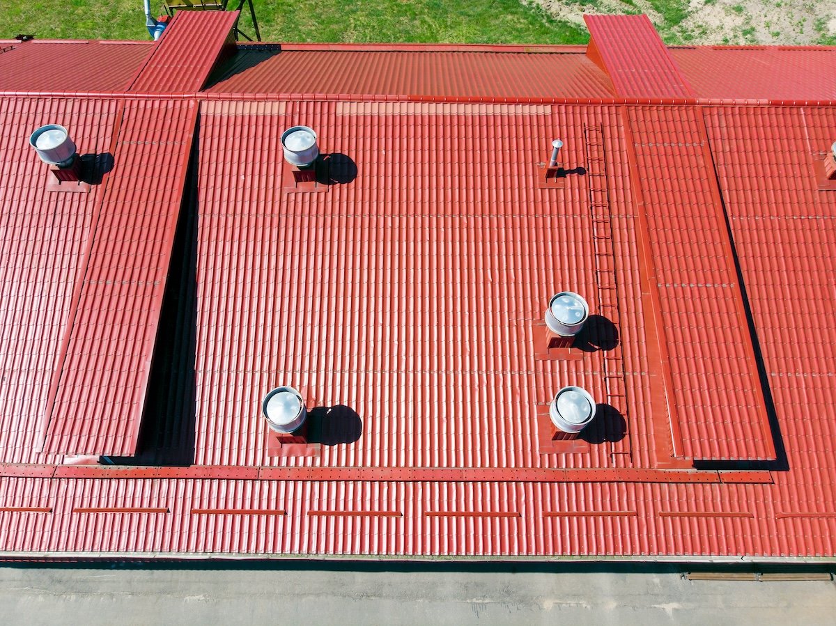 garner nc commercial Aerial view of a large red metal roof with multiple vents evenly spaced, nestled in a lush forest. The roof, adorned with vertical ridges, casts shadows on its surface while lying adjacent to a serene lake encircled by verdant greenery.