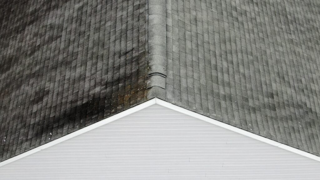 Aerial view of a roof with gray shingles and noticeable aging or moss on the left side. The roof forms a symmetric peak, with a white siding at the bottom. The right side of the roof appears cleaner and less weathered.