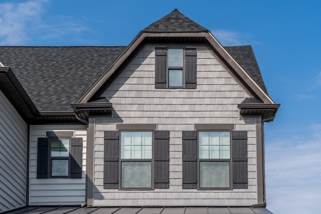 wake forest nc A two-story house with gray siding and black shutters showcases a steep roof under a clear blue sky. The second floor has three windows, and the architecture features gable elements with wooden shingles, highlighting the elegant roofing apex.