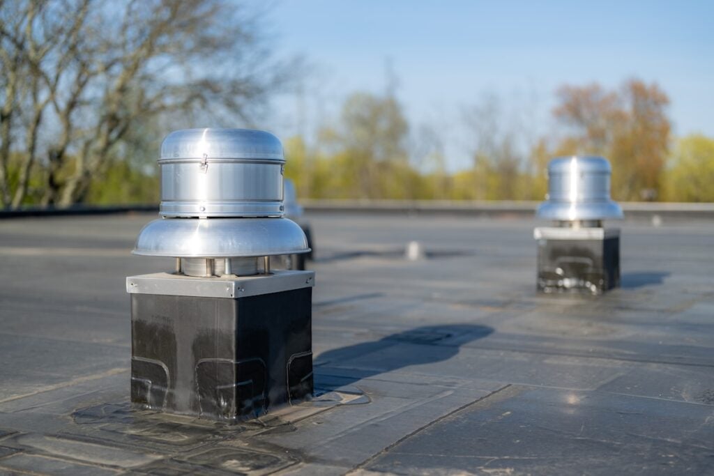 apex nc commercial Two silver metallic vents grace the flat rooftop, surrounded by a clear blue sky and trees reminiscent of a serene lake forest. The vents, mounted on sturdy black bases, glint as they capture sunlight in this bright and clear scene.