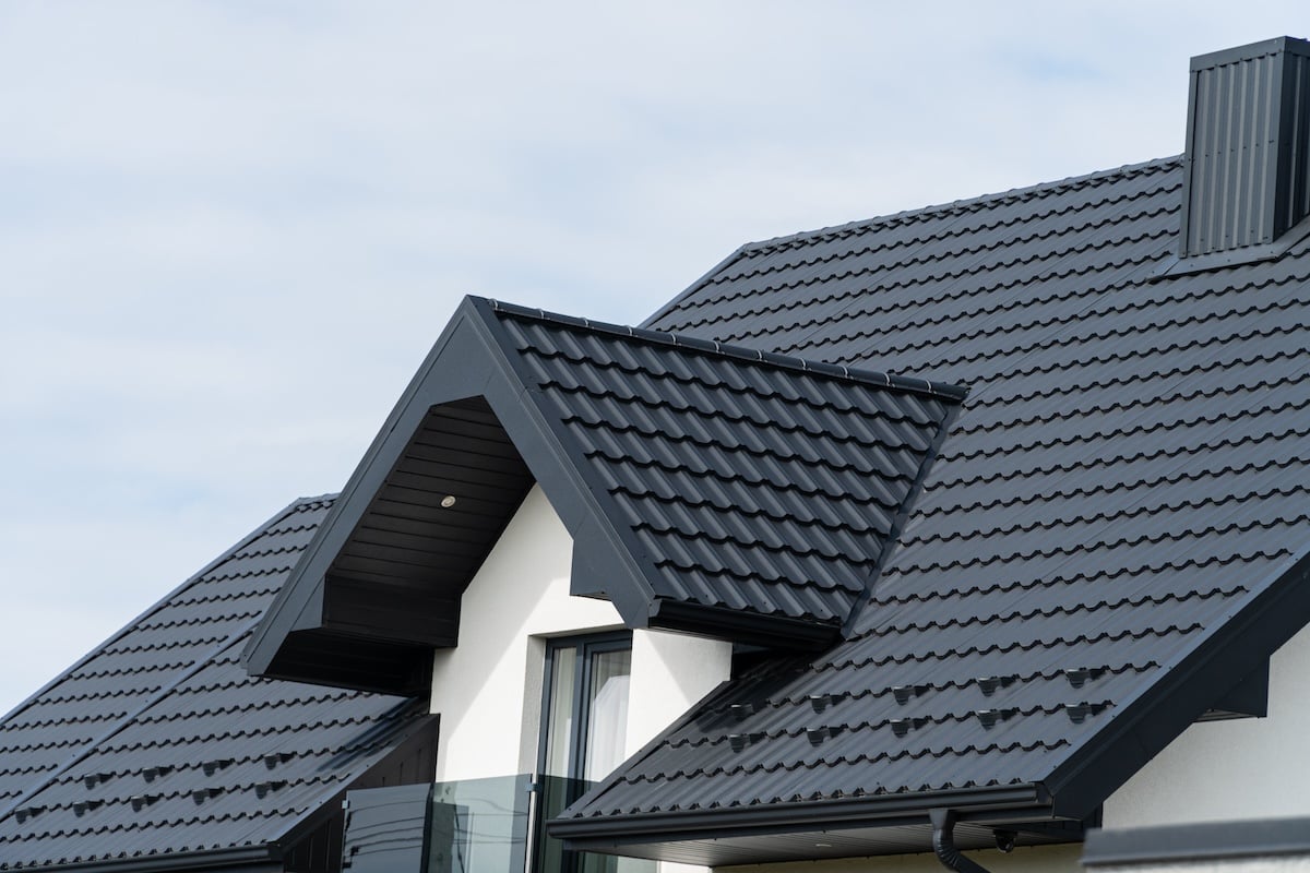 wake forest nc A modern house with a dark gray, textured metal roof by Roofing Morrisville, featuring gabled sections and a chimney. The building's exterior is light-colored, complementing the roof design. The sky is cloudy in the background.
