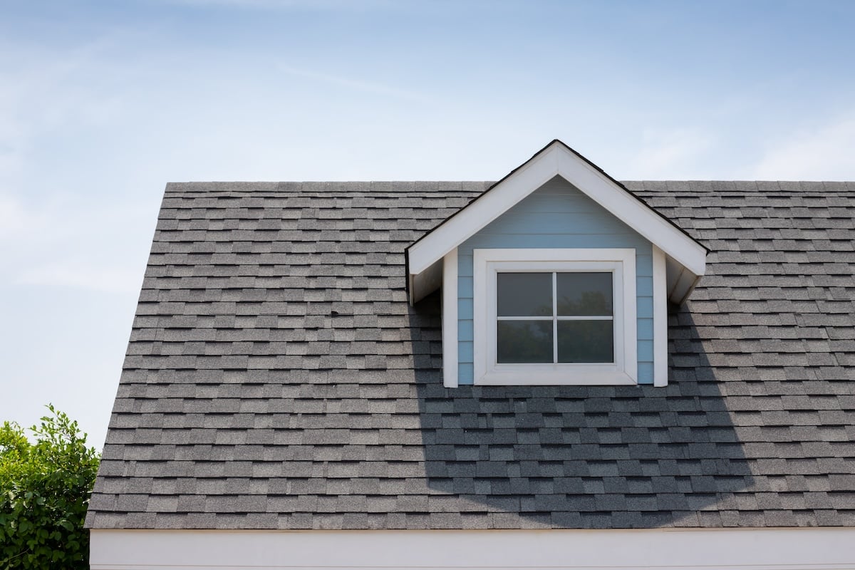 Chapel hill nc A gray shingled roof peaks gracefully at the roofing apex, featuring a dormer window painted light blue with white trim. Set against a clear blue sky, the scene is complemented by some green foliage partially visible on the left side.