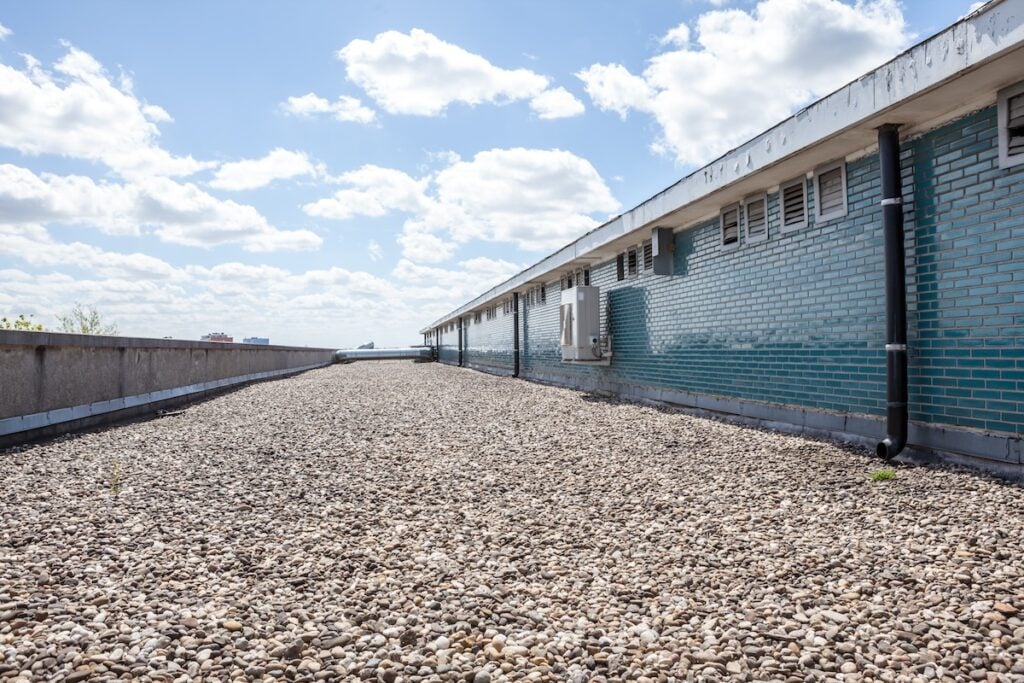 garner nc commercial The flat rooftop, reminiscent of a serene lake, is covered with gravel and supports a long building adorned with teal brick walls and multiple small windows. The sky is bright and scattered with clouds, while a distant forest seems to emerge on the horizon.