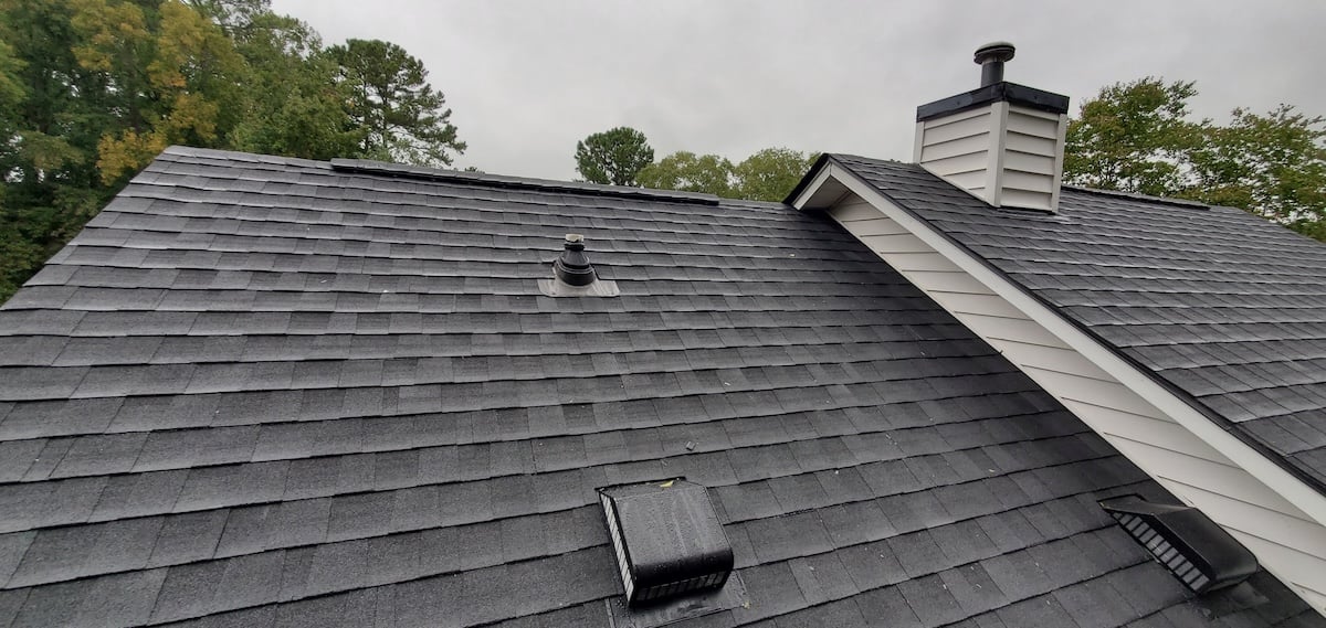 A gray shingle roof with multiple vents and a chimney stands prominently against a cloudy sky, epitomizing the quality craftsmanship of roofing in Durham. Trees with lush green foliage add a serene backdrop to this architectural display. cary nc