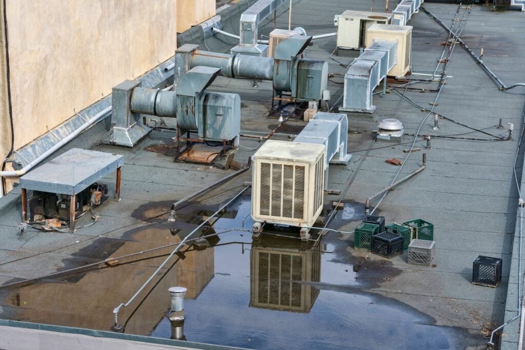Rooftop with various HVAC units and ductwork, surrounded by puddles of water, electrical cables, and scattered milk crates. The surface is flat and with signs of wear, alongside vent pipes and a low wall.