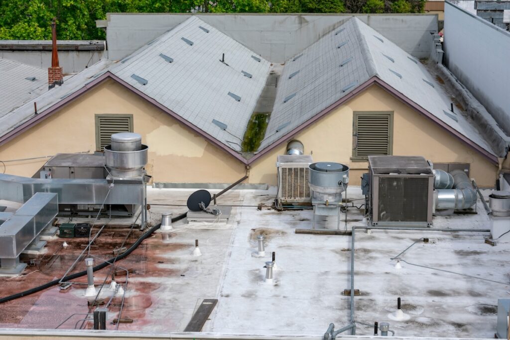 Rooftop with gray shingles featuring several utility units, including air conditioning and ventilation systems. A satellite dish and various pipes and ducts are spread across the flat concrete surface. Trees are visible in the background.