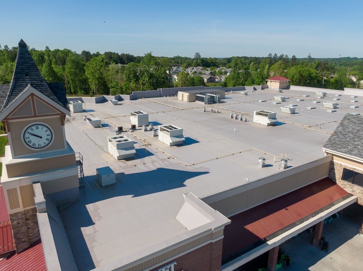 A flat rooftop of a commercial building in Lake Forest is dotted with several HVAC units and vents. The building, featuring a clock tower, is surrounded by lush trees and residential houses, all under a clear blue sky cary nc commercial