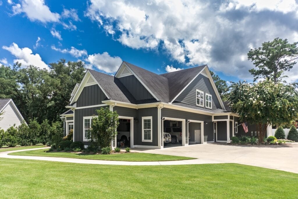 A modern, dark gray house with white trim and a steep roofing apex stands under a blue sky with clouds. It features a three-car garage with doors open, a curved driveway, and a well-maintained lawn surrounded by trees and bushes. An American flag hangs near the entrance. morrisville nc