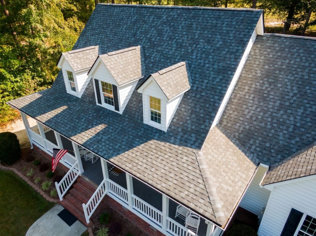Aerial view of a house with a grey shingle roof, showcasing three dormer windows. The porch, enhanced by white railings and an American flag, adds charm. Surrounded by trees and a neatly trimmed lawn, this roofing gem in apex nc stands out beautifully.