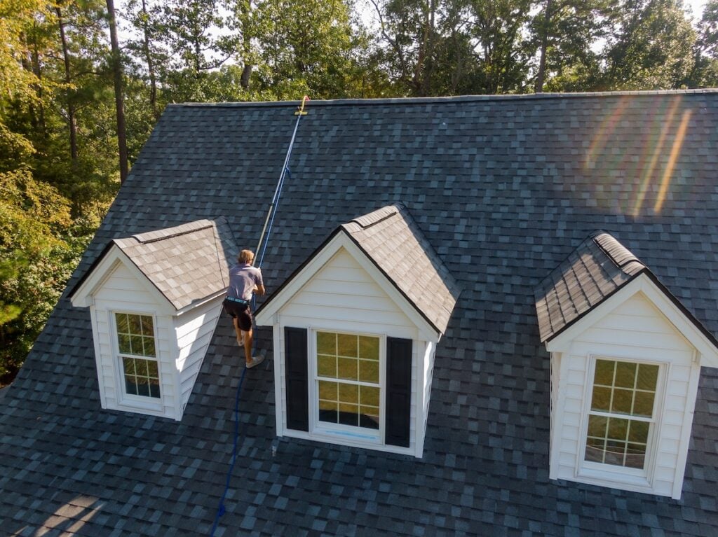 chapel hill nc A person meticulously cleans the gray shingled roof near the roofing apex using a long extendable pole. The roof boasts three dormer windows adorned with white siding and black shutters. Sunlight filters through trees in the background, enhancing the natural, bright setting.