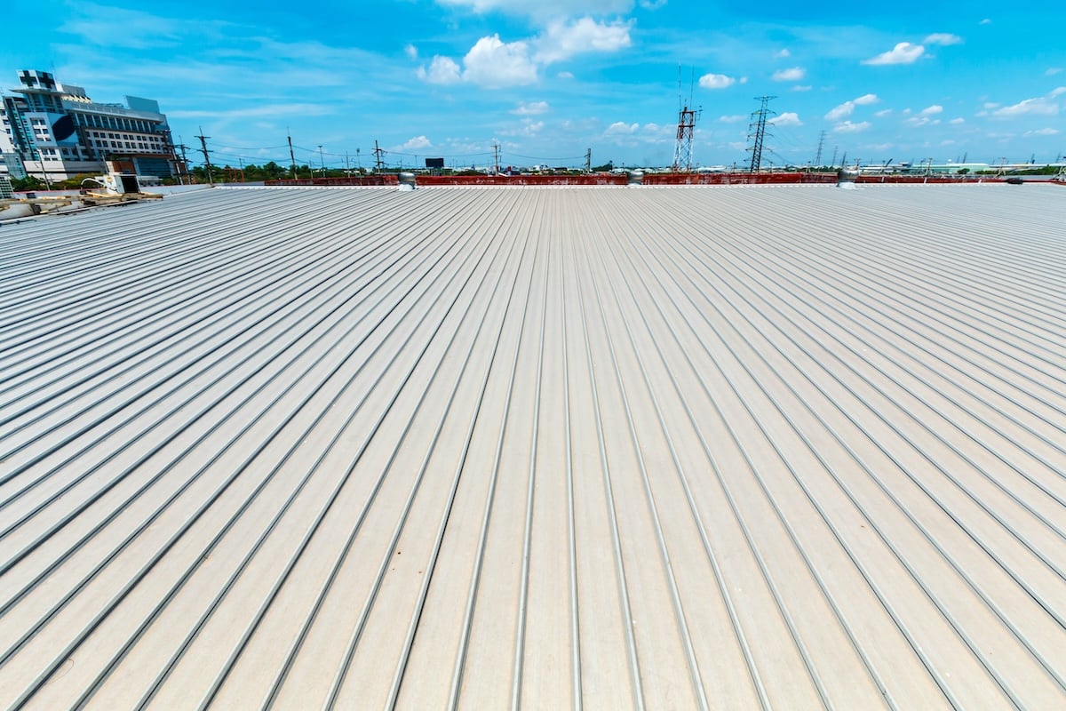 Wide-angle view of a large, flat industrial metal roof in Lake Forest, stretching under a bright blue sky with scattered clouds. In the background, buildings, electrical towers, and greenery beautifully frame the horizon. wake forest nc