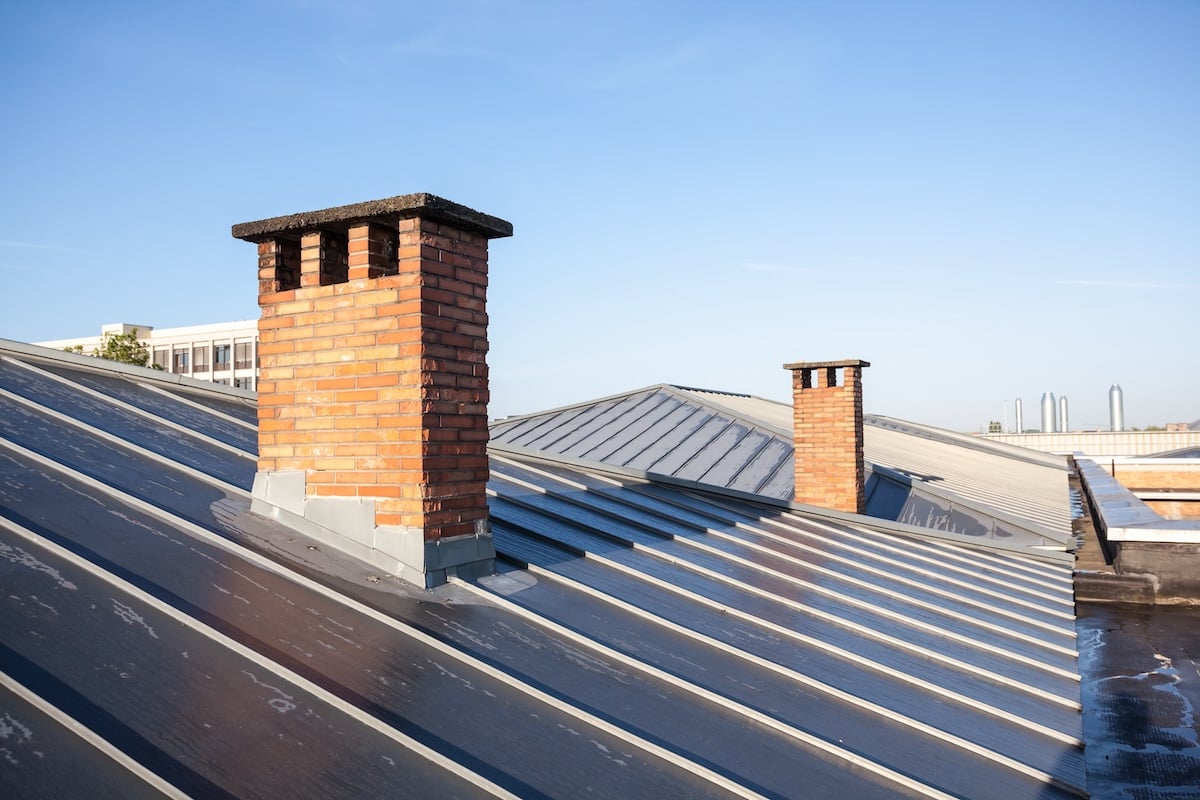A rooftop with two brick chimneys against a clear blue sky, reminiscent of serene mornings by Lake Forest. The metal panels gleam in the sunlight, while additional structures and rooftops create a harmonious backdrop. chapel hill nc