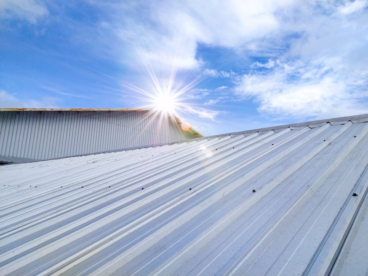 The image shows a metal roof in morrisville nc, with a bright sunburst at its edge against a partly cloudy blue sky. The sunlight highlights the ridges and texture of the corrugated metal surface.