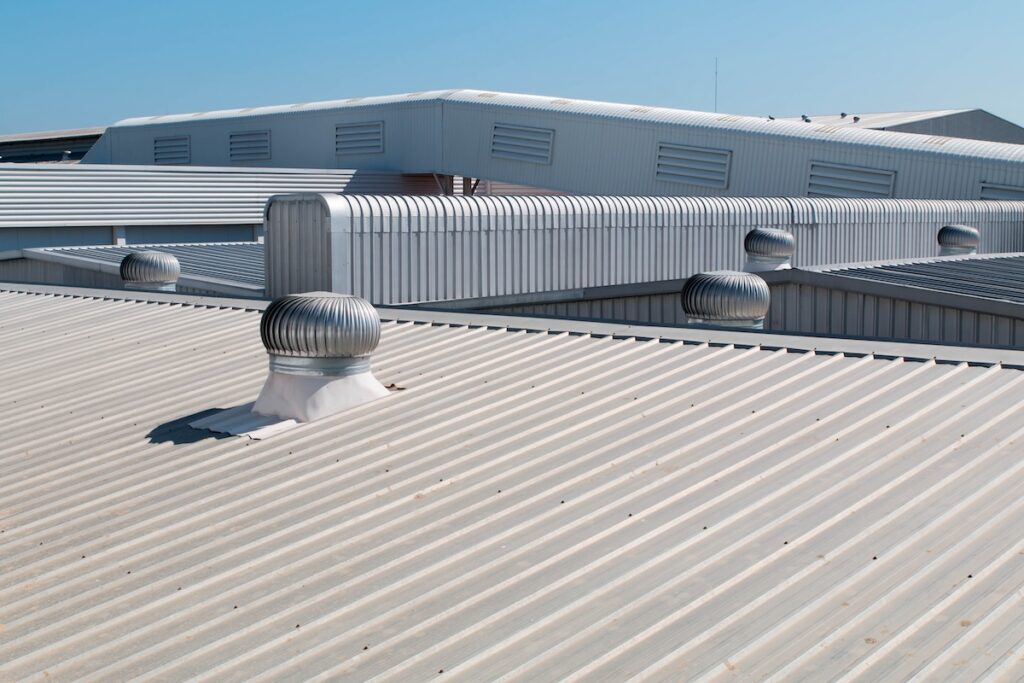 A rooftop in chapel hill nc features corrugated metal sheets with several cylindrical ventilators. In the backdrop, more rooftops with a similar structure stand out against the clear blue sky, showcasing the town's unique roofing charm.