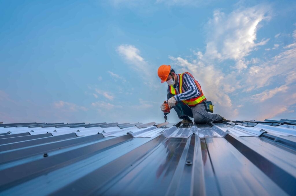 cary nc commercial A construction worker in an orange helmet and safety vest uses a drill on a metal roof in Lake Forest. Kneeling, surrounded by a clear blue sky with scattered clouds, the scene offers a low perspective that emphasizes the roof's metallic texture and reflection.