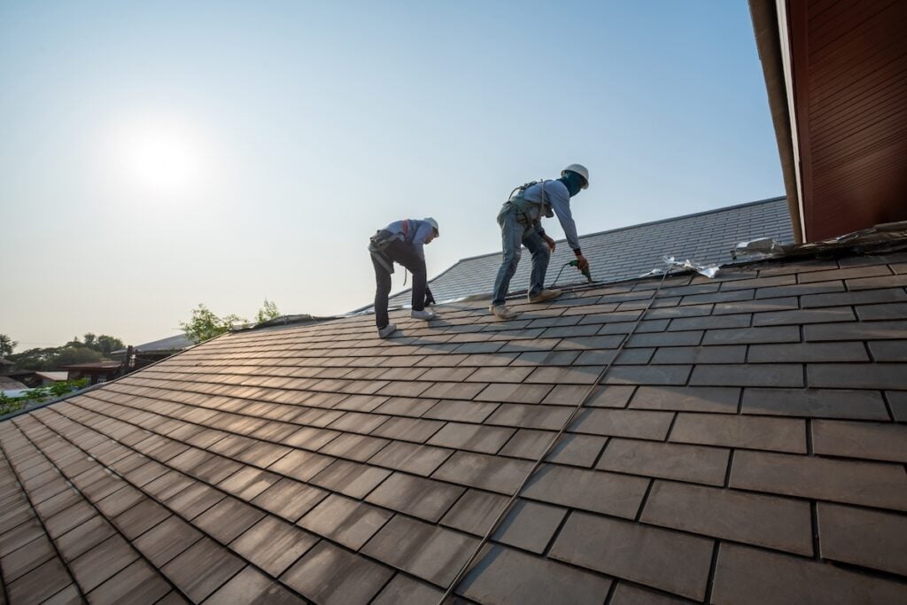 cary nc commercial Two workers wearing safety harnesses are installing tiles on a sloped roof under a clear sky in Lake Forest. The sun is low in the background, casting a warm glow over the residential area with trees visible in the distance.