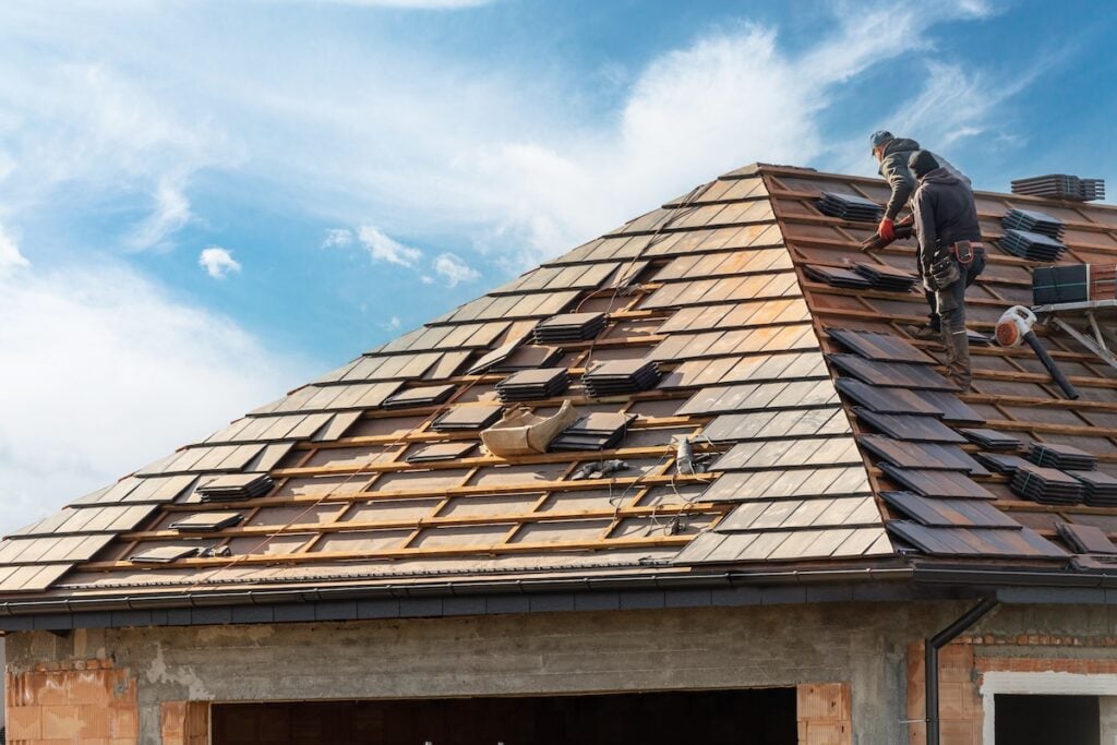 A worker installs shingles on a sloped roof of a house under construction in Durham. The sky is partly cloudy, and the roof structure is partially covered with wooden planks and roofing materials. A stack of shingles is visible nearby. cary nc