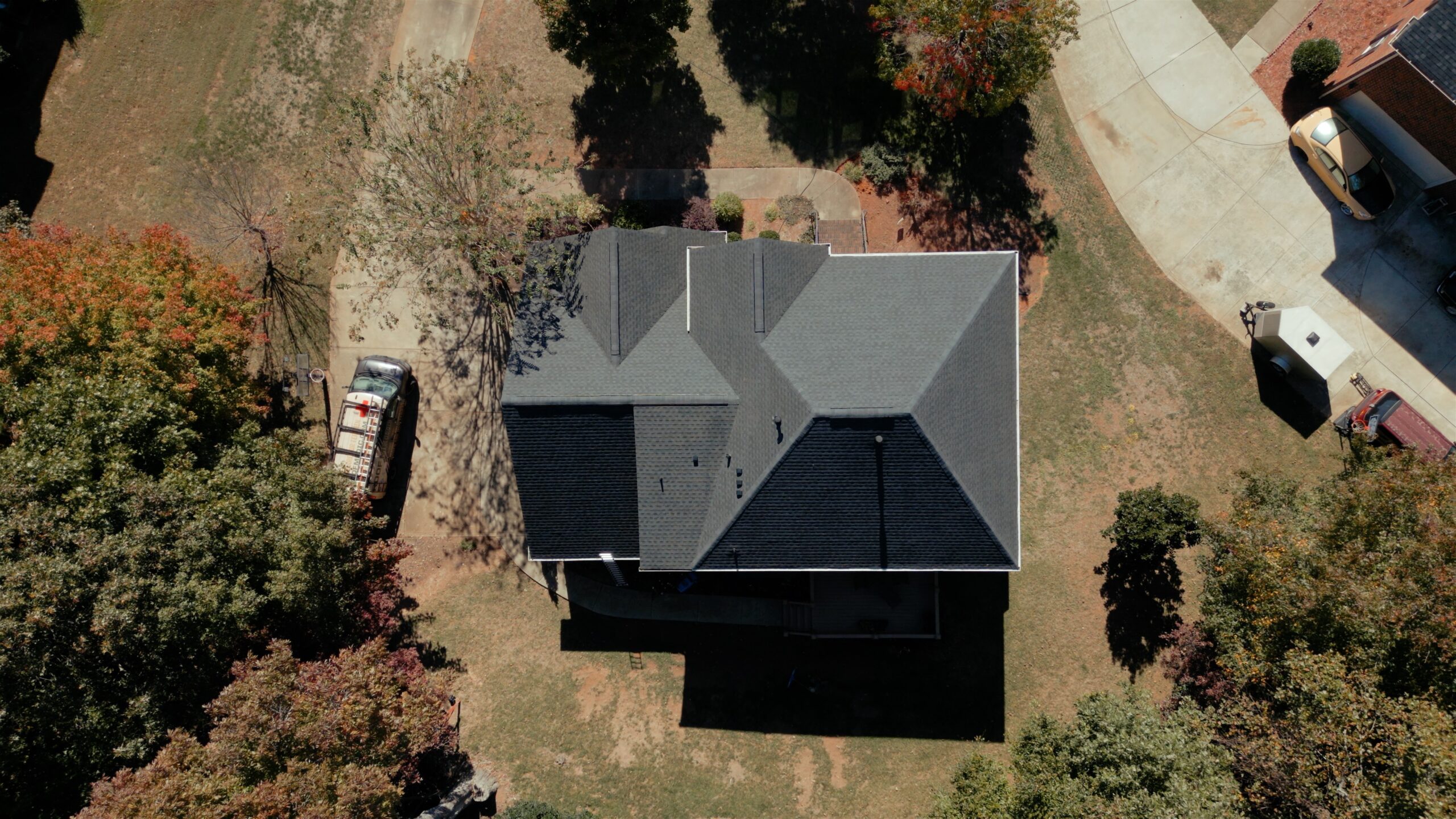 Aerial view of a house with a dark gray roof surrounded by grass and trees. The well-maintained roof suggests recent cleaning. A driveway to the left features a parked vehicle. Early autumn foliage adds color, while paved paths lead to a nearby curved road.